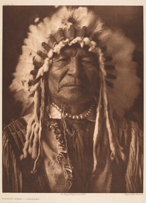 A sepia-toned portrait photograph of an Indigenous man wearing a feathered headdress, a necklace, and a tunic with fringe at the shoulders and breast.
