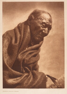 A sepia-toned portrait photograph of an older, balding Indigenous man wrapped in a dark blanket.
