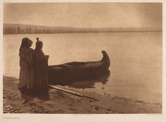 A sepia-toned photograph of three Indigenous people standing on a shore next to a canoe on calm water.