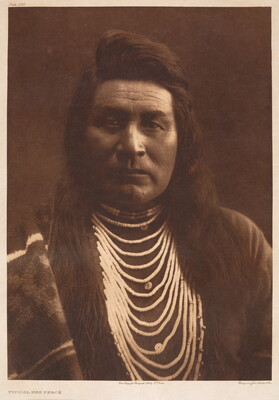 A sepia-toned portrait photograph of an Indigenous man, long hair hanging loose, wearing layers of beaded necklaces and a blanket over one shoulder.