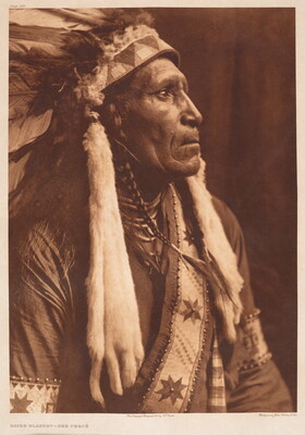 A sepia-toned portrait photograph of an Indigenous man in profile, wearing a feathered headdress with animal pelts hanging from the sides, beaded necklaces and a sash over a tunic.