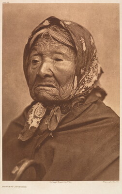 A sepia-toned portrait photograph of an elderly Indigenous woman with a deeply wrinkled face, wearing a kerchief over her hair, and a dark blanket or shawl around her shoulders.