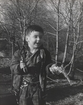 A black-and-white photograph of a White boy playing with two toy revolvers in a wooded setting.