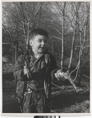 A black-and-white photograph of a White boy playing with two toy revolvers in a wooded setting.