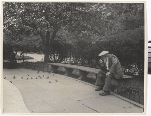 A black-and-white photograph of an older White man with a beard sitting on a stone bench in a park.