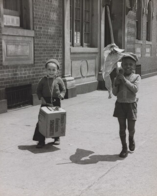 A black-and-white photograph of a White child banging a box like a drum and a Black child waving a flag of crumpled paper as they march on a city sidewalk.
