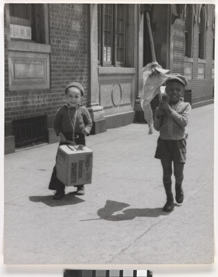 A black-and-white photograph a White child and one Black child smiling and marching on a city sidewalk, banging a box like a drum and waving a flag of crumpled paper.