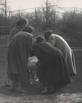 A black-and-white photograph of four women, backs to the viewer, bending down to look in a baby buggy.