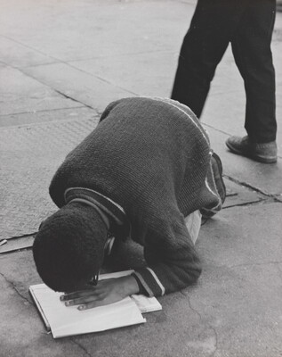 A black-and-white photograph of a Black boy hunched over his knees on pavement, his hand on a book and face close to his hand.
