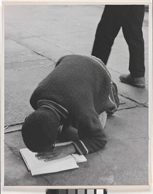 A black-and-white photograph of a Black boy hunched over his knees on pavement, his hand on a book and face close to his hand.
