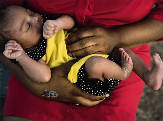 A color photograph of a dark-skinned sleeping baby in the arms of a Black woman wearing red clothing.