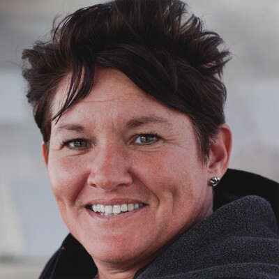 A color photograph of a smiling White woman with dimples, short, dark hair, earrings, wearing what looks like a hoodie.