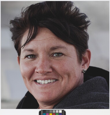 A color photograph of a smiling White woman with dimples, short, dark hair, earrings, wearing what looks like a hoodie.