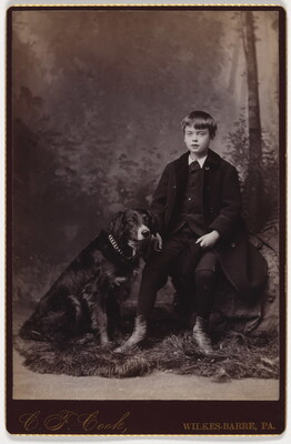 A black-and-white studio photograph of a young White boy seated next to a black dog.