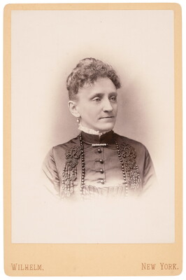 A sepia-toned portrait photograph of a White woman, curly hair pulled into an up-do, wearing a high-necked, dark beaded dress.