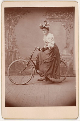 A black-and-white studio photograph of a White woman wearing a floral hat, white blouse, and long skirt posing on a bicycle in front of a scenic backdrop.