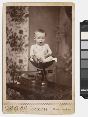 A sepia-toned studio photograph of a White baby wearing a necklace sitting in a scale.