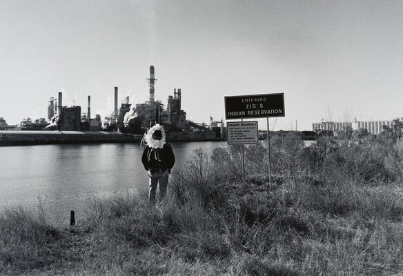 A black-and-white photograph of a man, face in shadow, wearing a feathered headdress standing in grass on the banks of a body of water next to a sign that reads, "Zig's Indian Reservation"; across the water is an industrial facility.