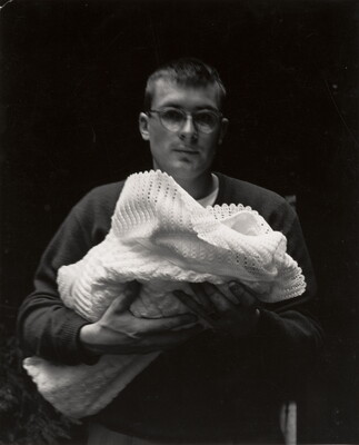 A black-and-white photograph of a White man wearing glasses holding a bundle of white blankets in front of him.