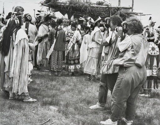 A black-and-white photograph of two women photographing a group of Indigenous people in cultural dress.