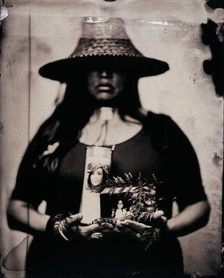 A black-and-white photograph of an Indigenous woman, eyes obscured by a straw hat, holding evergreen leaves, a candle, and photographs in her arms.