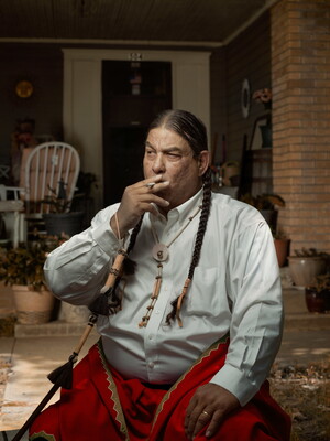 A color photograph of a seated Native American man with two, long braids smoking a cigarette in front of a porch.