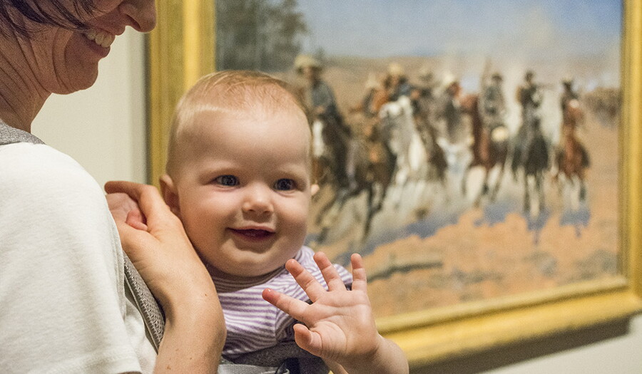 A woman holds a smiling baby who waves in a Carter gallery.