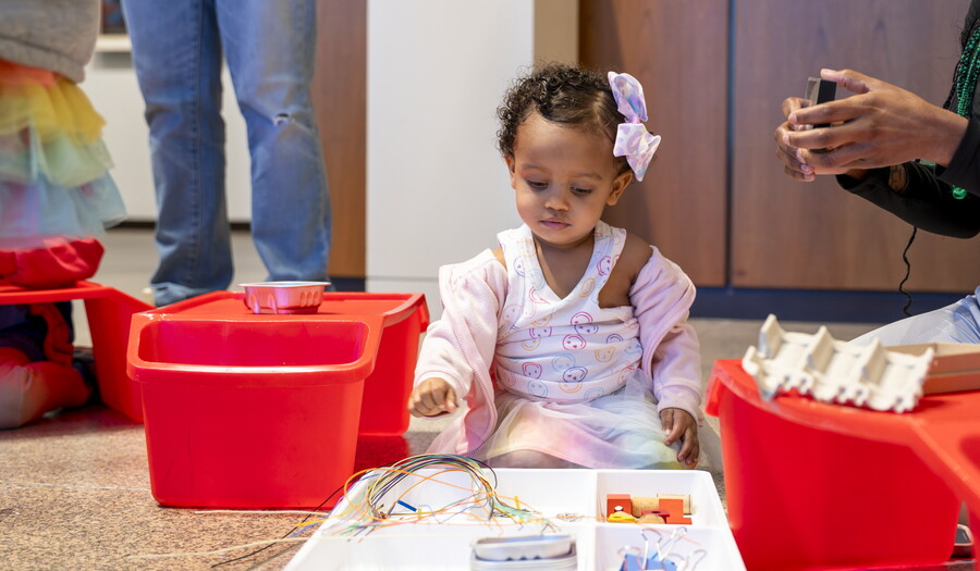 A young Black child with a giant pink bow in her hair plays with art supplies on the floor of a Carter gallery.