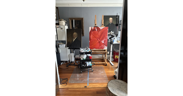 A painting of a red flower sits on an easel in a studio filled with other paintings.