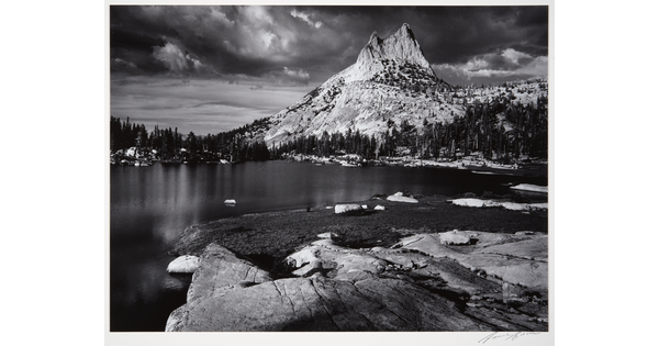 A black-and-white photograph of a rocky mountain peak from across a still lake surrounded by trees.