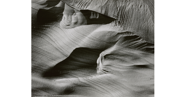 An abstract black-and-white photograph of sand dunes with horizontal and vertical striations that make it seem to be in motion.