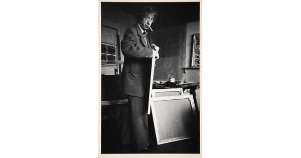 A black-and-white photograph of an older White man with a cigarette in his mouth holding a canvas in an artist's studio.