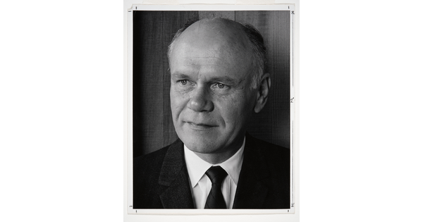 A black-and-white portrait photograph of a mostly bald White man gazing to the side wearing a dark jacket, dark tie, and white shirt.