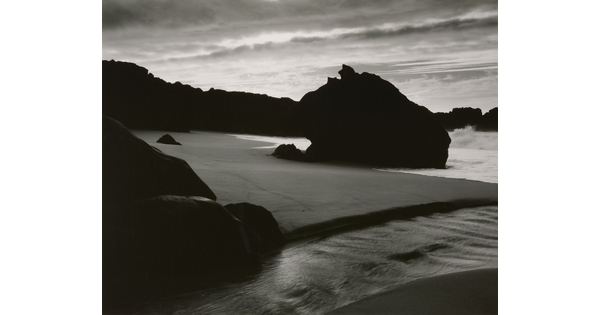 A black-and-white photograph of the sand and sea next to a shore covered with large rocks.