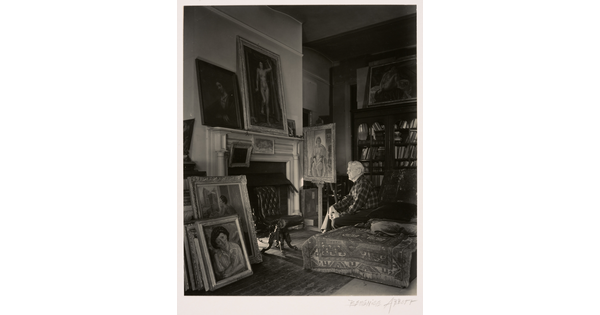 A black-and-white photograph of an older White man seated on a divan near a fireplace; elaborately framed paintings lean above the mantel, against the wall, and on an easel.