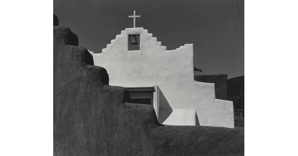 A black-and-white photograph of a terraced adobe bell tower with a cross on top, partially hidden by a terraced adobe wall.