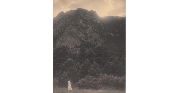 A black-and-white photograph of a woman in a white dress standing at the base of a rocky mountain covered with trees.