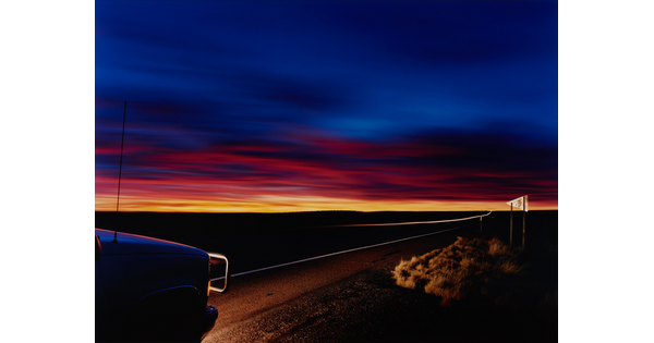 A color photograph of a deep blue sky over a line of orange and yellow sunset in a flat landscape; the lights from the front of a truck in the left corner illuminate part of the road.