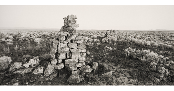A black-and-white photograph of several tower-like mounds of stones, receding into the distance, in an arid landscape dotted with sagebrush.