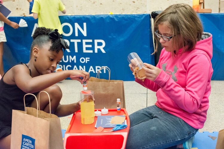Two children are kneeling on either side of a small plastic red workbench during an art-making activity.