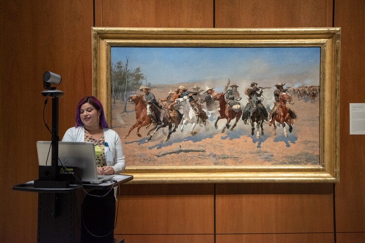 A woman is standing behind a podium equipped with distance learning equipment, including a camera and laptop. The podium is positioned in front of and to the left of a large painting.