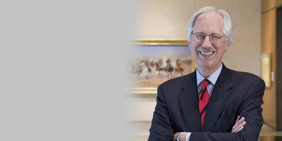 Man with gray hair and mustache, blue eyes behind gold-rimmed glasses stands with his arms crossed in a blue sports jacket and a red and black striped tie.