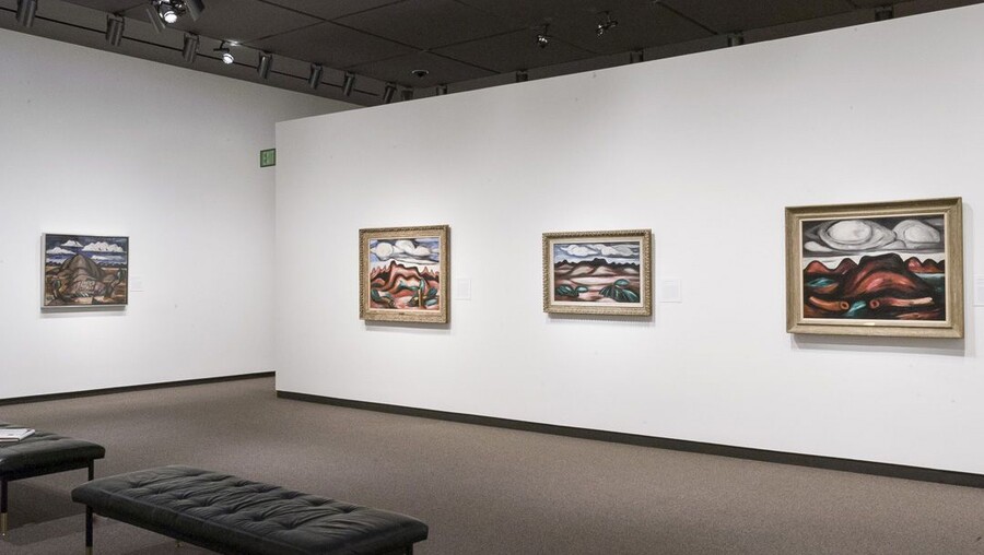View of gallery with white walls; four framed abstract landscape paintings hang on the walls.