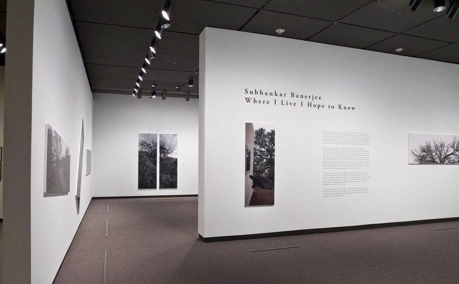 View of exhibition title wall featuring two large-scale photos mounted on a white wall; other photos can be seen on walls within the exhibition.