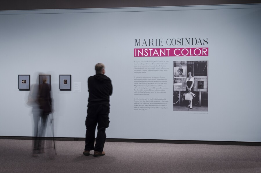 View of exhibition title wall with two people standing in front of it. On the wall are three framed photographs, the exhibition text, and an enlarged black-and-white photo of the artist.