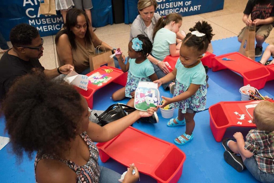 Adults and children sit on a blue mat on the floor with red lap desks in front of them on which they are working on art projects.