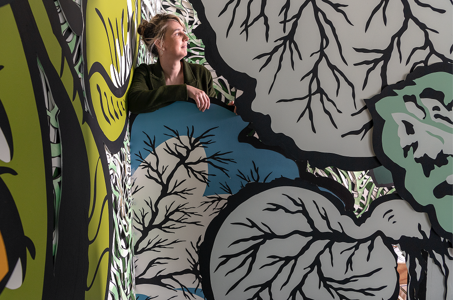 A blonde woman with her hair pulled up on her head looks off to her left from behind large-scale, cartoon-like botanical illustrations