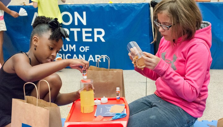 Two children are kneeling on either side of a small plastic red workbench during an art-making activity.