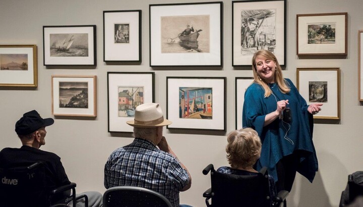 A smiling woman with long blonde hair wearing a turquoise-colored shawl stands in front of a wall of framed art; in front of her is a row of elderly people, two of whom are sitting in wheelchairs, listening to her talk