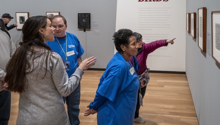 Four people stand in a gallery; (L-R) A woman with long dark hair and her back to the camera gestures with her right hand toward art on the wall; in front of her is an adult in a blue t-shirt looking at her; to the woman's right, an older woman with short hair wearing a blue t-shirt leans in toward art on the wall to get a better look; behind the second woman is a person wearing a red jacket with their left arm outstretched pointing toward art on the wall